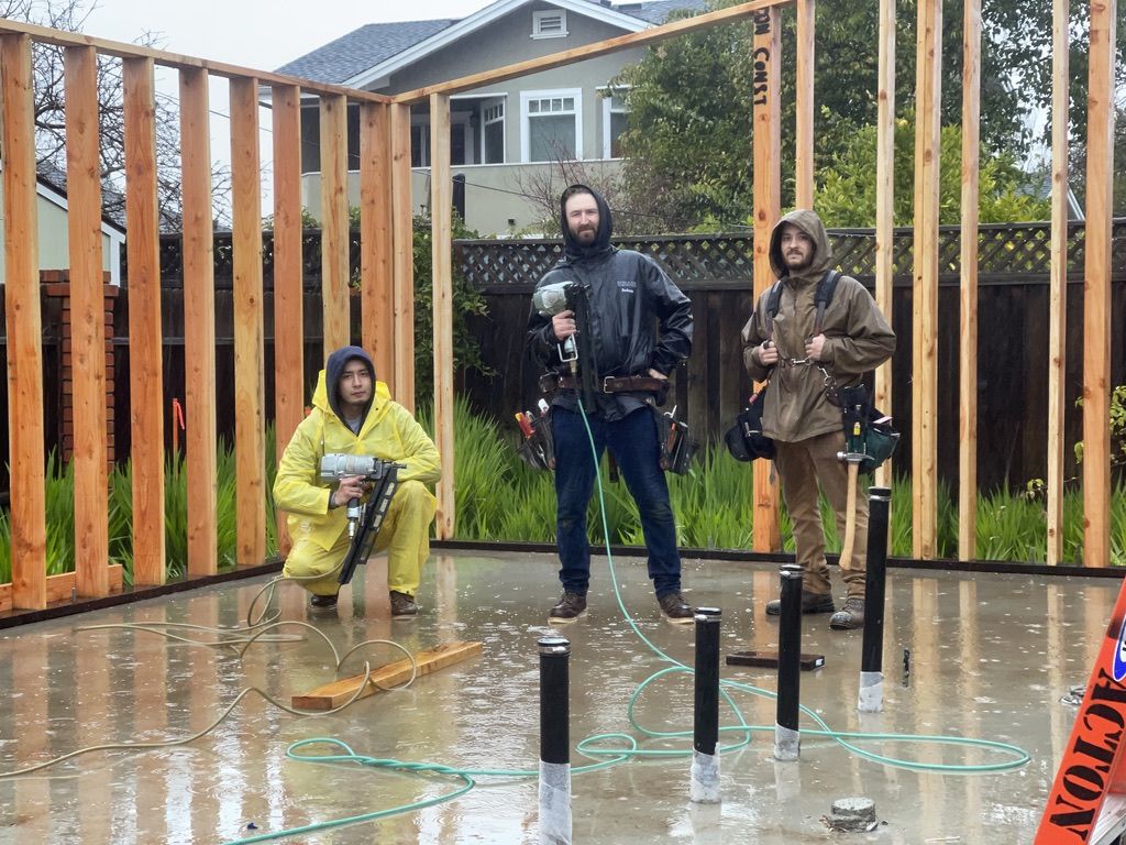 Three framers in rain gear working on a partially framed wall on a wet slab in heavy rain