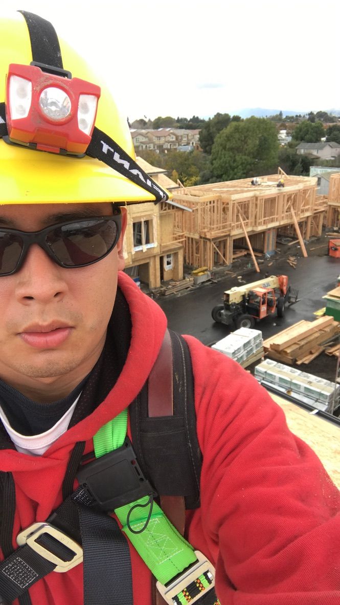 Tied off in a fall-protection harness on a multifamily framing site, with a telehandler and stacked lumber below